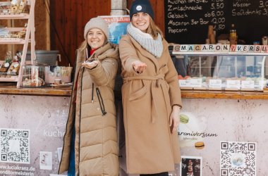 Zwei Frauen stehen vor ihrem Verkaufsstand, lächeln in die Kamera und präsentieren Ihre Macarons 
